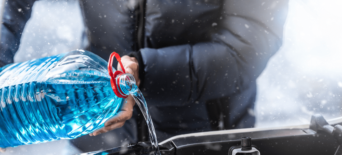 person in a navy down jacket pouring blue liquid from a clear bottle with a red lid into a car engine