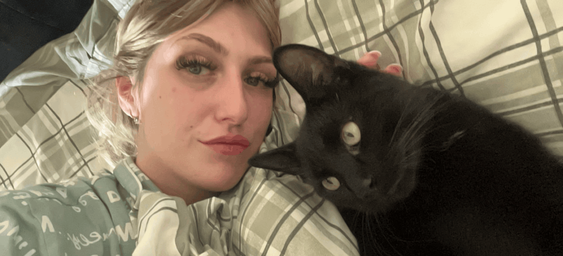 Shannon Brady lies beside Hammy, a black cat, on a bed as they look towards the camera.